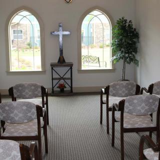 A small chapel interior with rows of chairs, a cross at the front, and arched windows.
