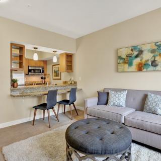 A modern living room with an open layout connected to the kitchen, featuring a comfortable sofa, stylish chairs, and abstract artwork on the wall.