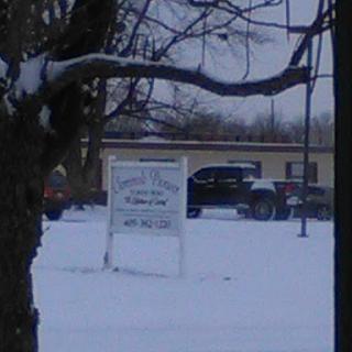 A snowy scene featuring a sign near a tree, with vehicles in the background.