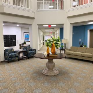 A spacious lobby area featuring comfortable seating, a central table with flower arrangements, and a well-lit hallway.