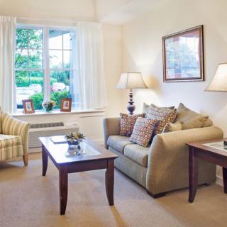 A cozy living room with a sofa, patterned armchair, and coffee table, featuring soft lighting and a large window with a scenic view.