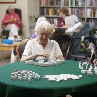An elderly woman sitting at a table in a library, playing a game of dominoes.