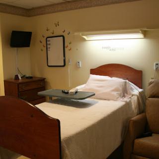 A hospital room featuring a bed with a tray table, a TV mounted on the wall, and a comfortable chair.