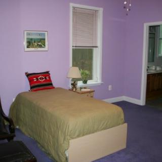 A cozy bedroom with purple walls, featuring a single bed and a side table with a lamp.