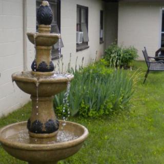 A three-tiered fountain in a garden beside a building, surrounded by lush greenery.
