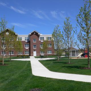 A brick building with a white pathway surrounded by greenery and trees under a clear blue sky.