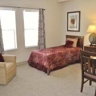 A cozy bedroom featuring a single bed with patterned bedding, a desk and chair, a lamp, and two windows with curtains.