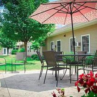 A peaceful patio setting with chairs and a table under a red umbrella, surrounded by lush greenery and flowers.