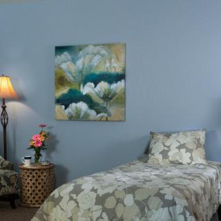 A cozy bedroom with a single bed, side table with flowers, an armchair, and a decorative painting on the wall.