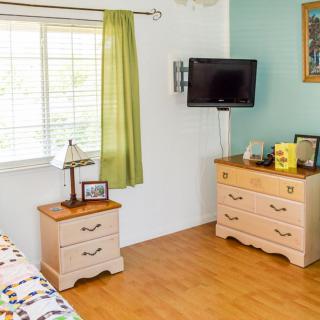 A bright, tidy bedroom featuring a dresser, wall-mounted TV, and a window with green curtains.