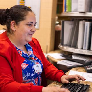 A woman in a red cardigan smiles while working at her desk, typing on a computer.
