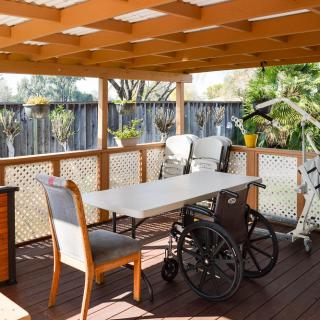 A sunny patio with a table, chairs, and a wheelchair under a wooden pergola.