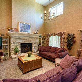 A cozy living room with a stone fireplace, purple sofas, and a wooden coffee table, adorned with decorative plants and bookshelves under a high ceiling.