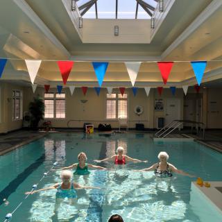 A group of seniors participate in a water exercise class in an indoor pool, illuminated by natural sunlight streaming through the skylight.