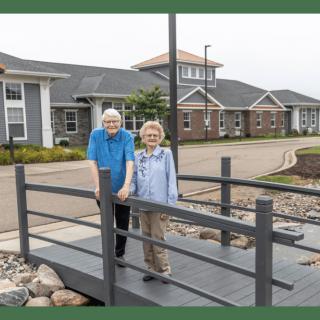 An elderly couple stands on a small bridge in front of a residential area.