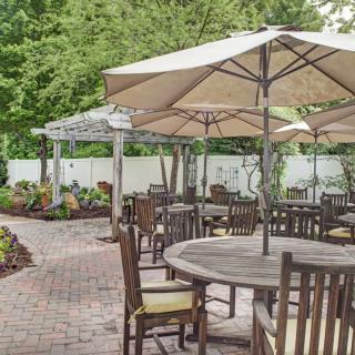 A tranquil outdoor patio with wooden tables and chairs shaded by large umbrellas, surrounded by lush greenery.