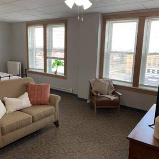A cozy living room with large windows providing natural light, featuring a sofa, armchair, TV, and indoor plant.