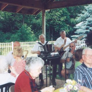 An outdoor social event with musicians playing an accordion and guitar under a gazebo, surrounded by elderly attendees.