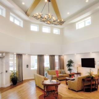 A spacious and elegantly designed living room featuring a high ceiling with a chandelier, modern furniture, and ample natural light from multiple windows.
