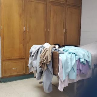 A bed cluttered with various clothes, set against a wooden wardrobe backdrop.