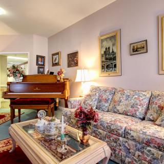 A cozy living room with a vintage-style decor, featuring a floral patterned sofa, a grand piano, and various paintings on the wall.