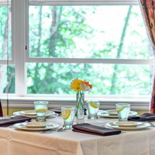 An elegantly set dining table with glasses, plates, and a flower centerpiece by large windows.