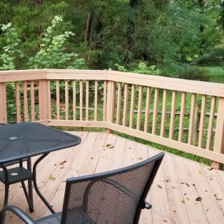 A wooden deck with a black metal table and chairs, surrounded by lush greenery.