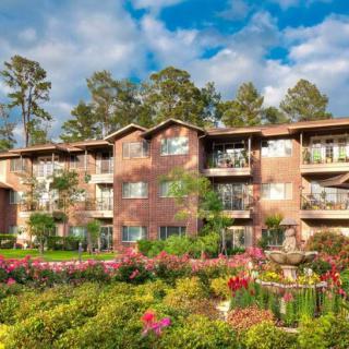 A scenic view of an apartment building surrounded by vibrant flowers and trees under a cloudy sky.