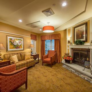 A cozy living room with warm lighting, featuring a fireplace, armchairs, and a sofa adorned with artwork on the walls.