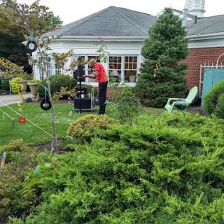 A man tends to the garden in front of a house, surrounded by lush greenery.