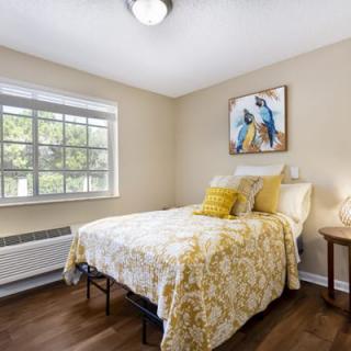 A cozy bedroom with a bed covered in decorative pillows and a floral comforter, featuring a window that lets in natural light, a piece of artwork above the bed, and a nightstand with a lamp.