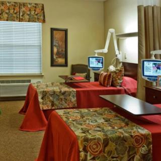 A comfortable and well-equipped nursing home room with two hospital beds, floral bed covers, and individual TVs for each bed.