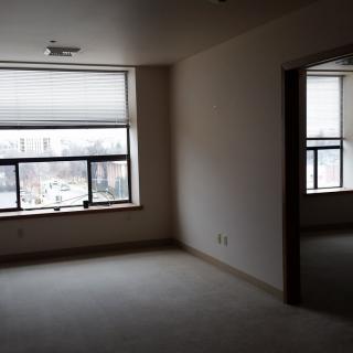 A vacant apartment room with large windows and closed blinds, featuring a carpeted floor.