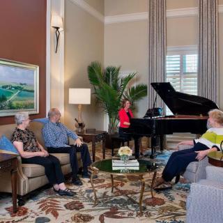 A cozy living room with people gathered around a grand piano, engaged in conversation.