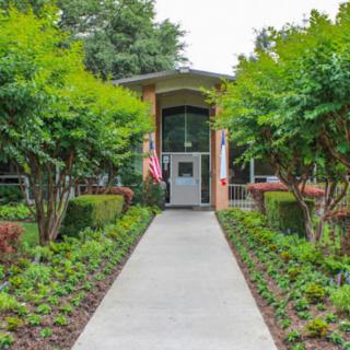A pathway lined with trees and shrubs leading to a building entrance adorned with a flag.
