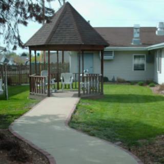 A backyard featuring a gazebo with chairs, located beside a house with a neatly trimmed lawn and a concrete pathway.
