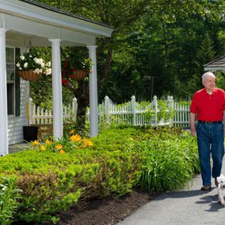 An elderly man walking his dog along a garden path in front of a house.