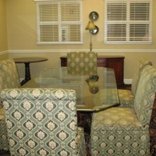 A dining room featuring a glass-topped table and patterned upholstered chairs against a backdrop of shuttered windows.