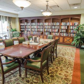A cozy library space featuring a large wooden table surrounded by chairs, with shelves filled with books in the background.