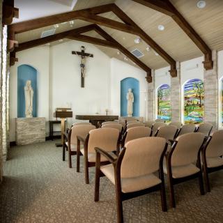 Interior view of a small chapel with seating, wooden beams, a crucifix, statues, and stained glass windows.