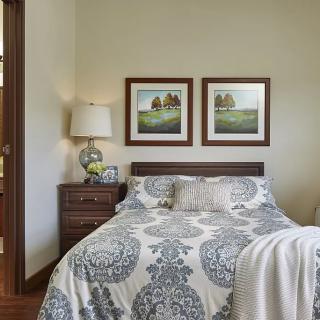 A cozy bedroom featuring a neatly made bed with decorative pillows and patterned bedding, flanked by two framed pieces of artwork on the wall and a bedside table with a lamp.