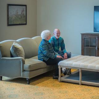 An elderly couple sitting together on a couch in a living room, engaged in conversation.