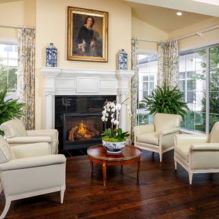 A cozy living room with a fireplace, surrounded by armchairs and decorated with plants and a portrait painting above the mantel.