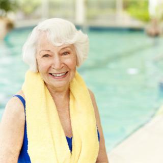 An elderly woman smiling by the poolside with a yellow towel around her neck.