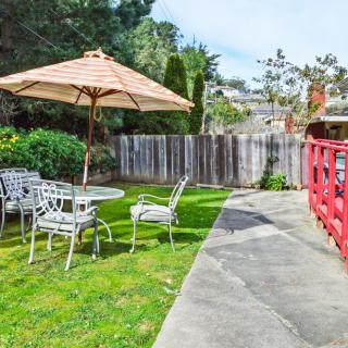 A cozy backyard patio with outdoor furniture and a large umbrella, surrounded by greenery and a wooden fence.