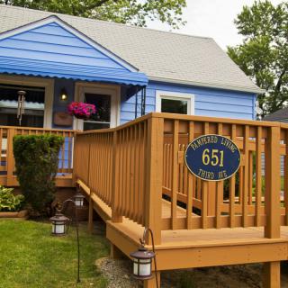 A cozy blue house with a wooden front porch and address sign, surrounded by a small garden.