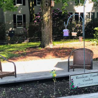 A serene garden area featuring comfortable chairs, a purple bird feeder, and a sign marking the space.