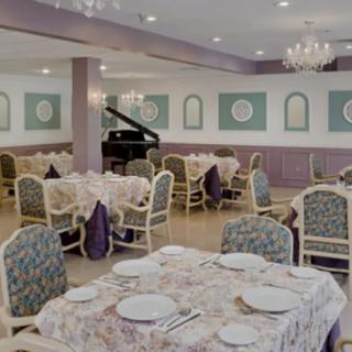 An elegant dining room featuring multiple tables with patterned tablecloths, a grand piano in the corner, and chandeliers overhead.