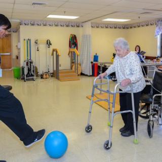 An elderly woman using a walker participates in an exercise activity with the help of a caregiver, while a blue ball is placed on the floor.