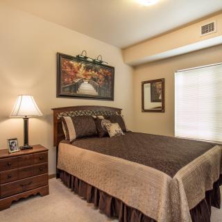 A cozy bedroom with warm lighting, featuring a neatly made bed, a wooden nightstand with a lamp, and artwork on the walls.